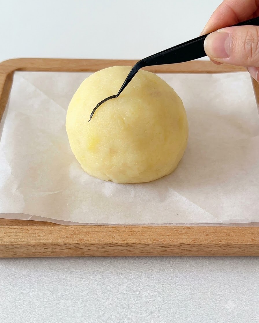 Une main tenant une fine pince noire plaçant un morceau incurvé dalgue nori sur une tête de pomme de terre sphérique.