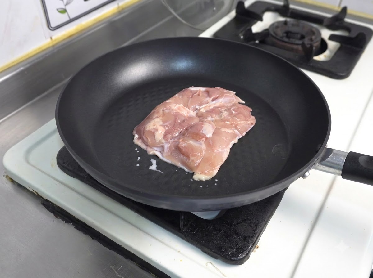 Un morceau de cuisse de poulet cru côté peau vers le bas dans une poêle antiadhésive sombre.