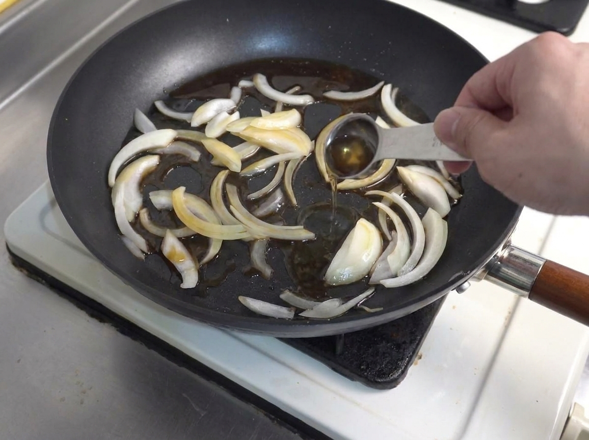 Une cuillère de mesure versant un assaisonnement liquide dans une poêle avec des tranches doignon sautées.