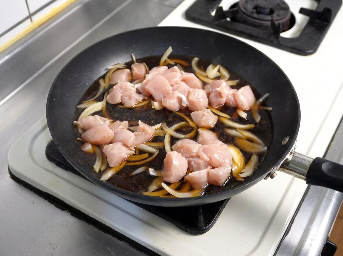 Des morceaux de poulet cru placés dans une sauce sombre frémissante avec des oignons à lintérieur dune poêle.