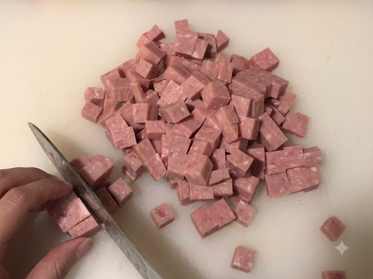 Du spam coupé en petits cubes sur une planche à découper blanche, avec des oignons et des oignons verts hachés en arrière-plan.