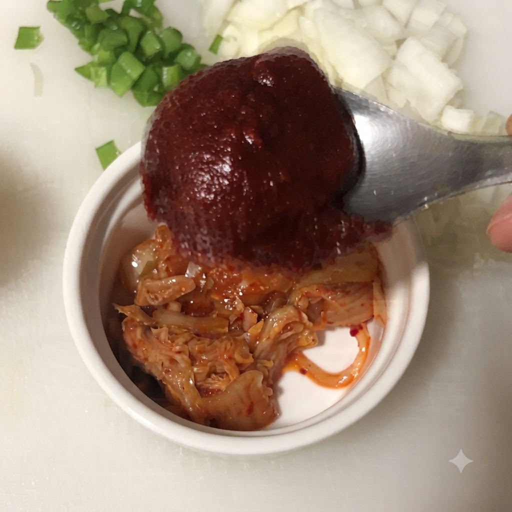 Une cuillère de pâte de piment coréenne tenue au-dessus dun bol contenant du kimchi haché.