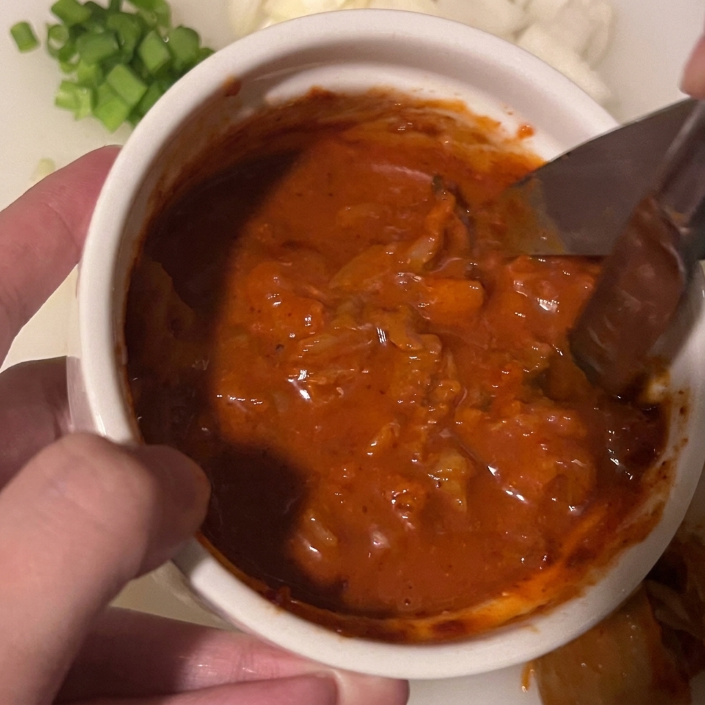 Des ciseaux de cuisine coupant des morceaux de kimchi baignant dans un mélange de sauce rouge épaisse dans un petit bol blanc.