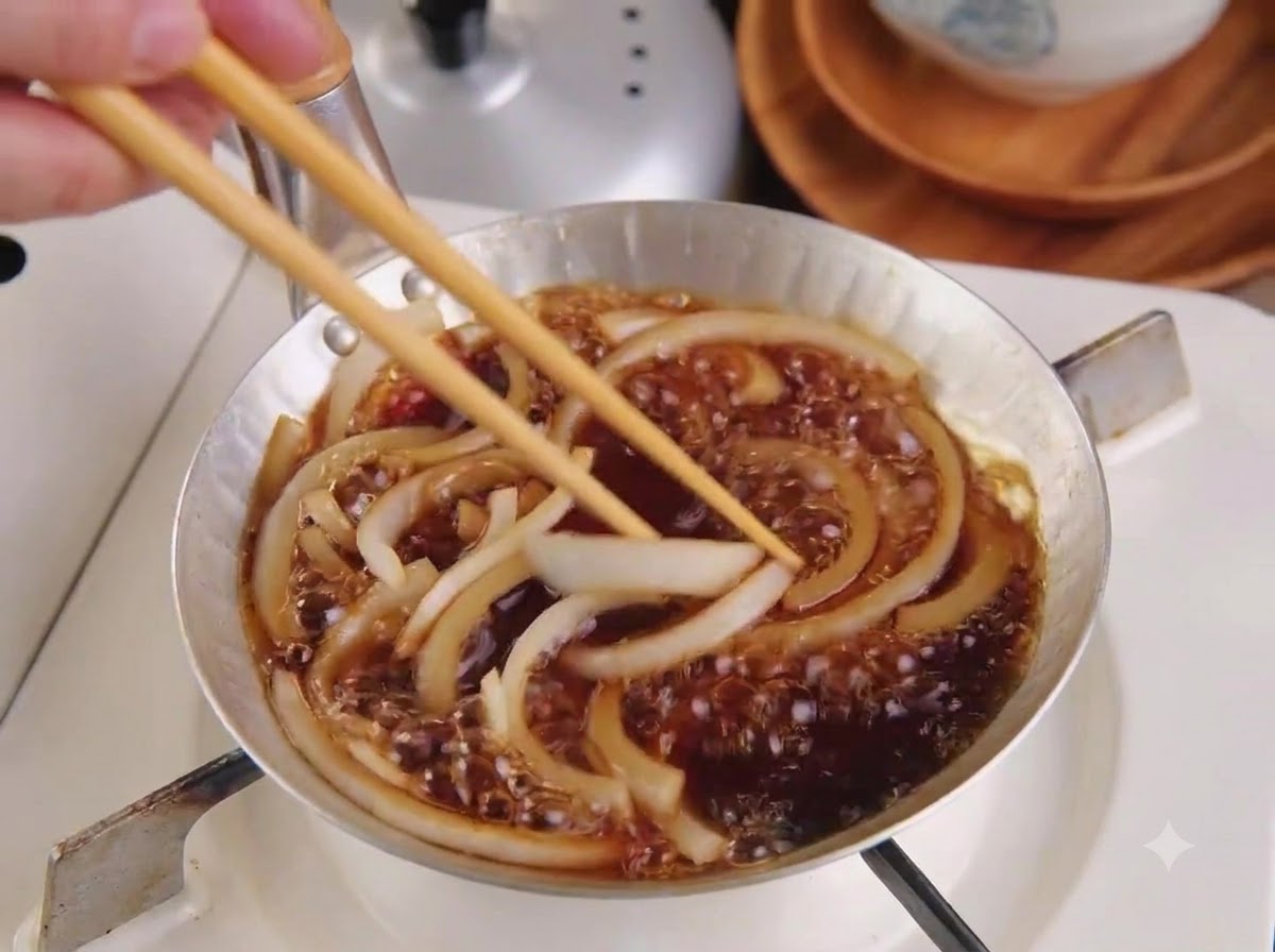 Une côtelette de porc tranchée disposée dans une poêle par-dessus les oignons et la sauce frémissants à laide de baguettes en bois.