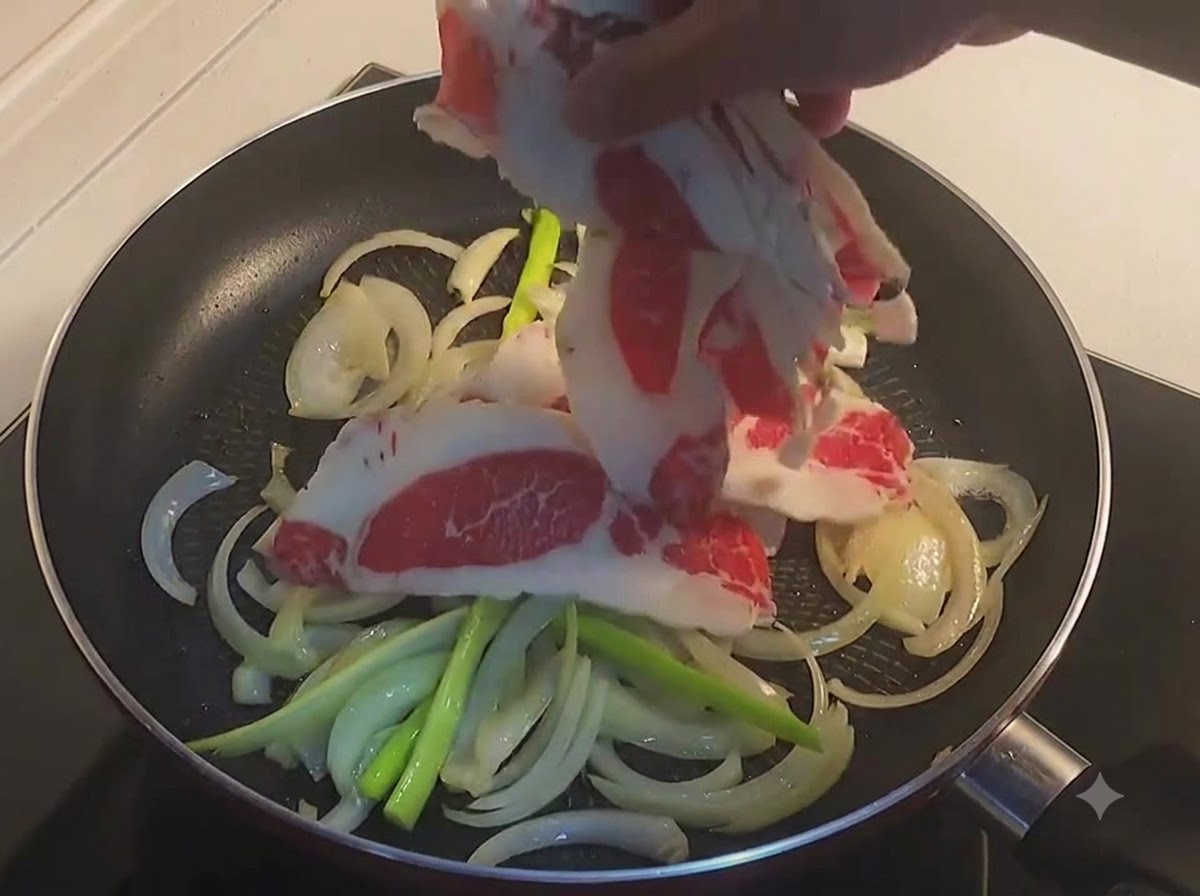 Une main plaçant de fines tranches de poitrine de bœuf crue dans une poêle avec des oignons tranchés et des oignons verts.