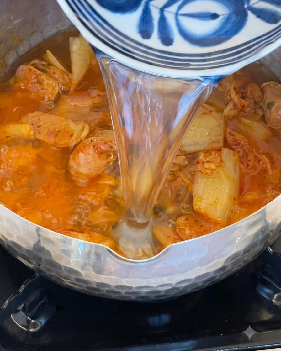 De leau versée depuis un bol bleu et blanc dans une casserole bouillonnante de kimchi et de saucisses.