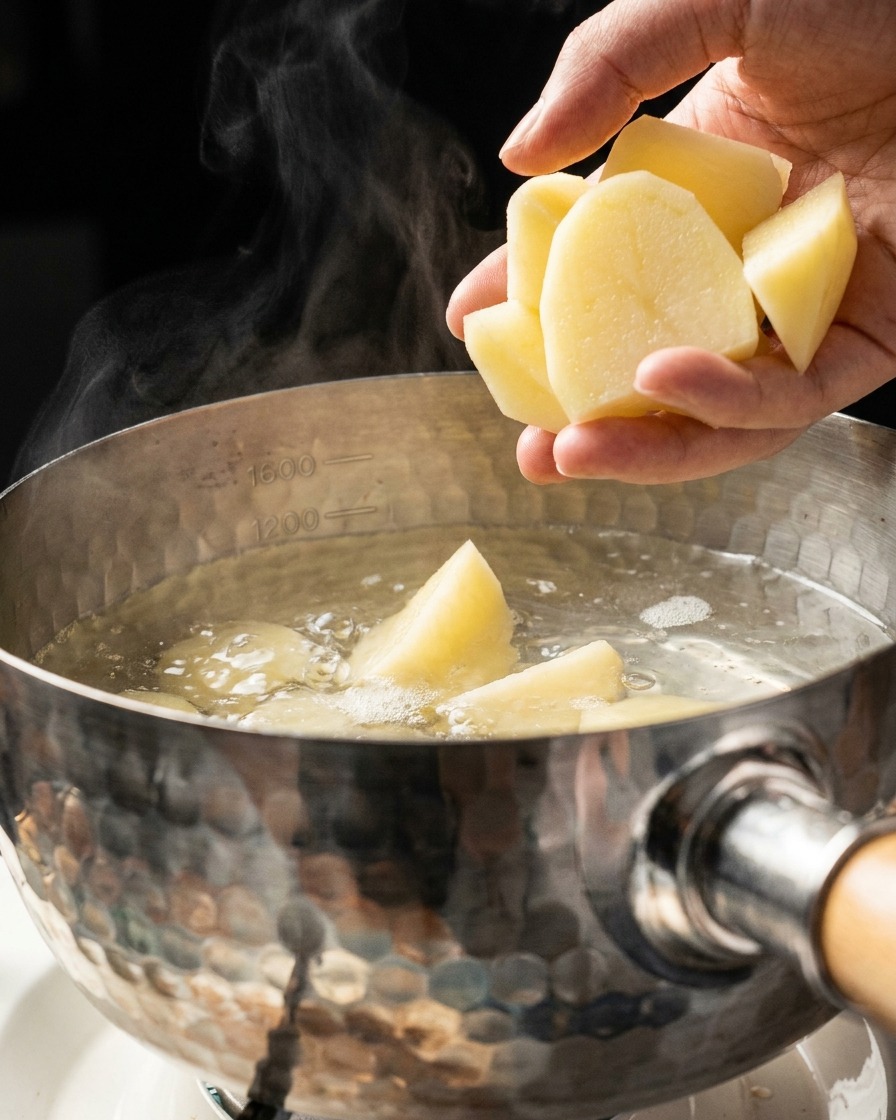 Une main jetant des morceaux de pommes de terre coupés dans une casserole en métal remplie deau bouillante et dégageant de la vapeur.