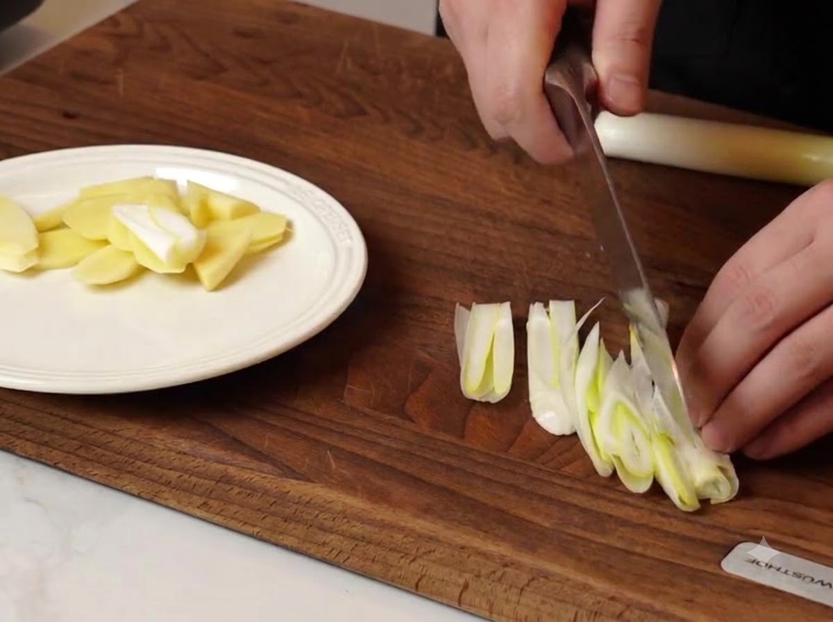 Un cuisinier coupant des oignons verts et des morceaux de gingembre sur une planche à découper en bois à côté dune assiette blanche.