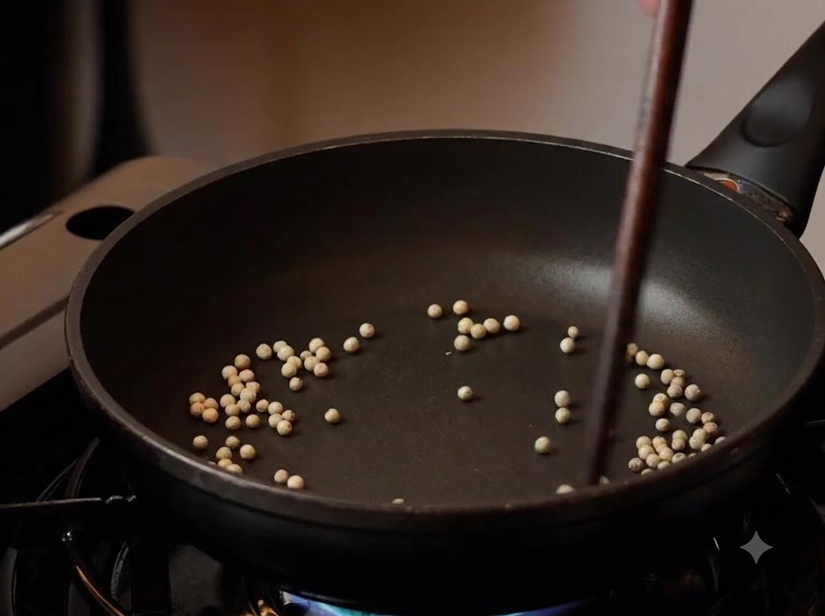 Des grains de poivre blanc torréfiés dans une poêle noire avec des baguettes les remuant à feu doux.