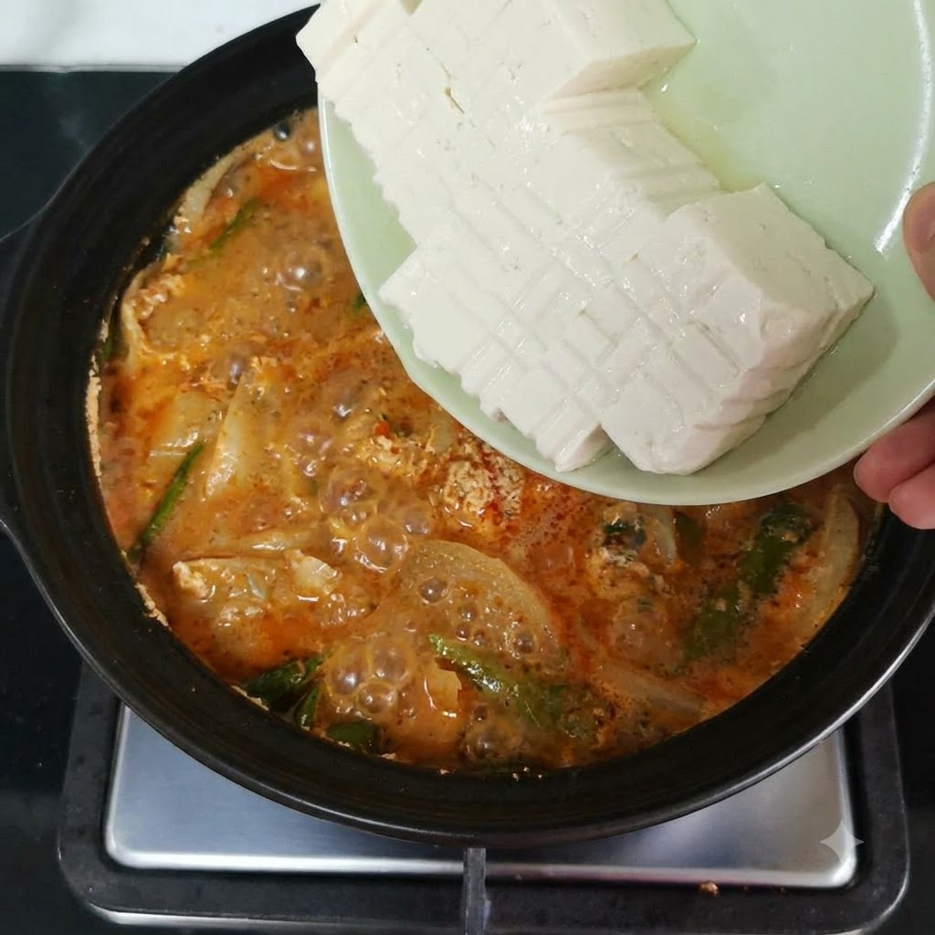 Des tranches de tofu soyeux basculées dune assiette dans une casserole frémissante remplie dun bouillon rouge-orangé et de légumes.
