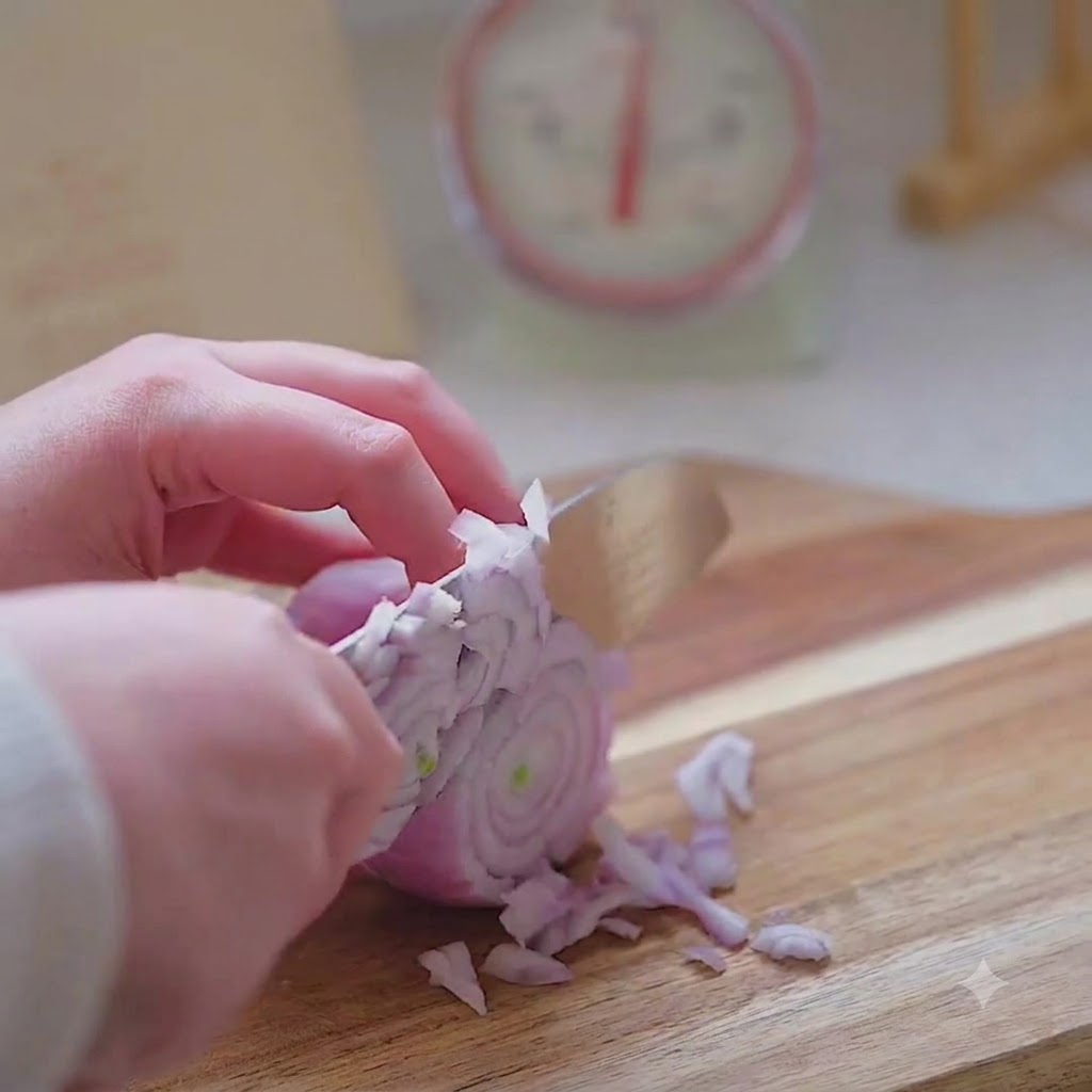 Une main utilisant un couteau de cuisine pour émincer finement un oignon rouge sur une planche en bois.