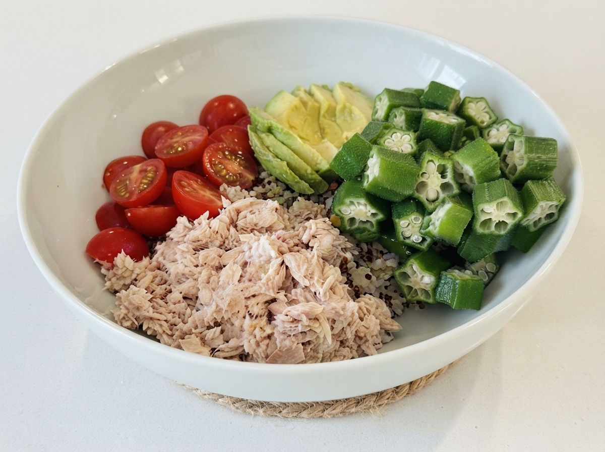 Un bol blanc rempli dune base de riz, garni de sections de thon, tomates cerises, avocat en tranches, et gombos.
