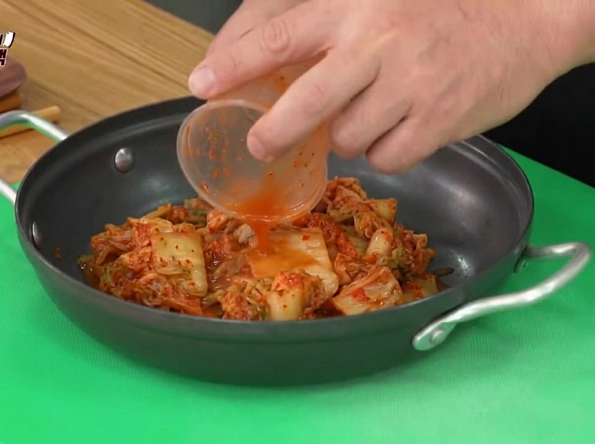 Du kimchi aigre ajouté dun gobelet en plastique dans une casserole de cuisson noire.