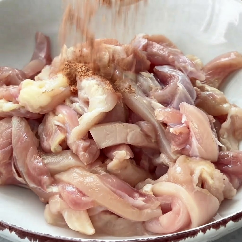 Des épices saupoudrées sur un bol rempli de lanières de poulet cru prêtes à être mélangées.