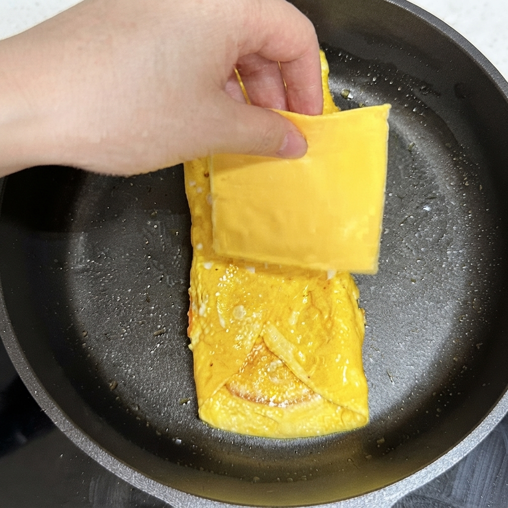 Une main plaçant une tranche carrée de fromage jaune sur lensemble dœufs cuits dans la poêle à frire.