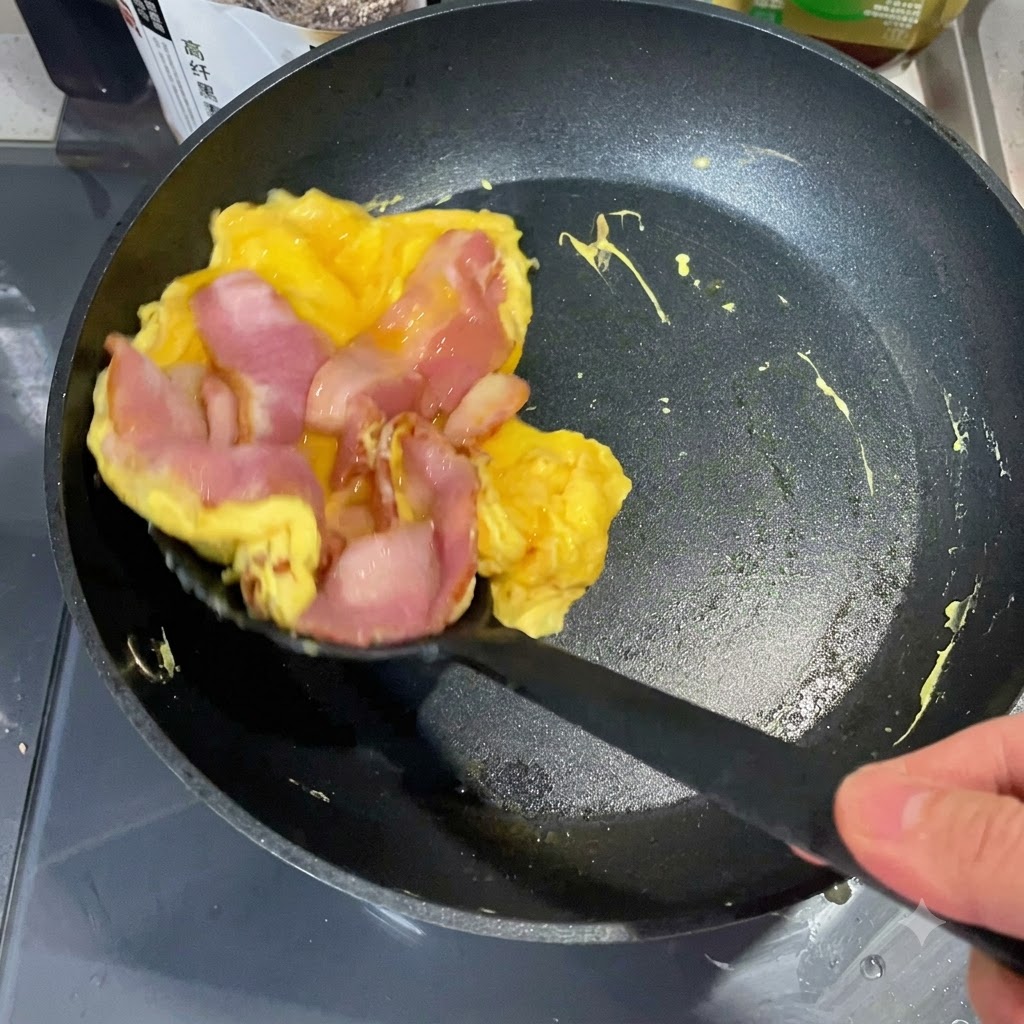 Une spatule soulevant une pile dœufs et de bacon cuits dune poêle pour la placer sur une moitié de muffin anglais toasté.
