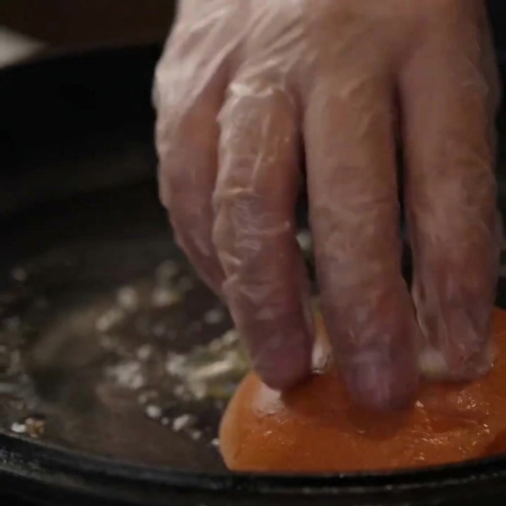 Une main gantée pressant des pains à burger dans une poêle en fonte beurrée pour les toaster.
