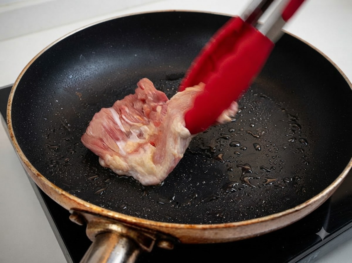 Un haut de cuisse de poulet cru en train dêtre placé dans une poêle chauffée avec de lhuile à laide de pinces rouges.