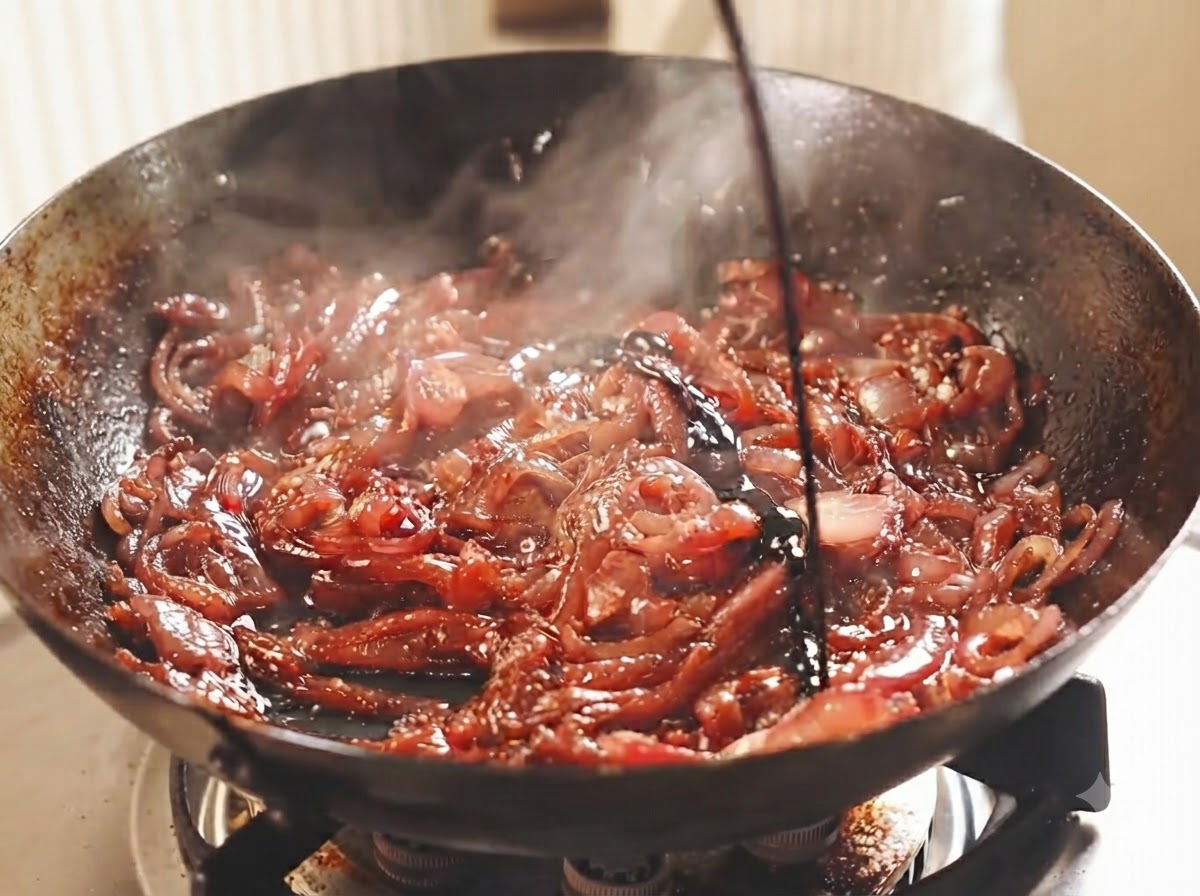 Des tranches doignons rouges en train de mijoter dans un wok sombre, arrosées de vin et de vinaigre.