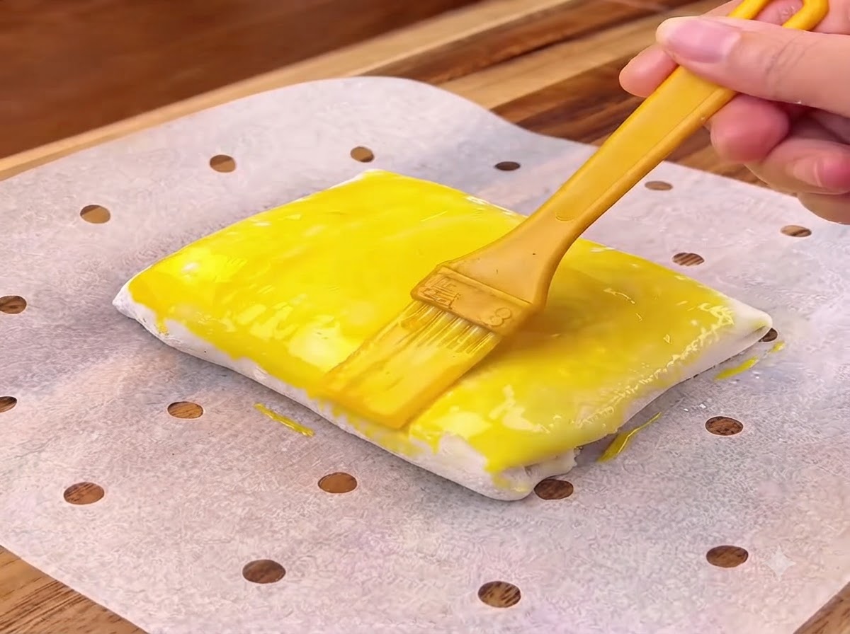 Une dorure à lœuf jaune appliquée au pinceau sur la surface dun carré de pâte pliée.