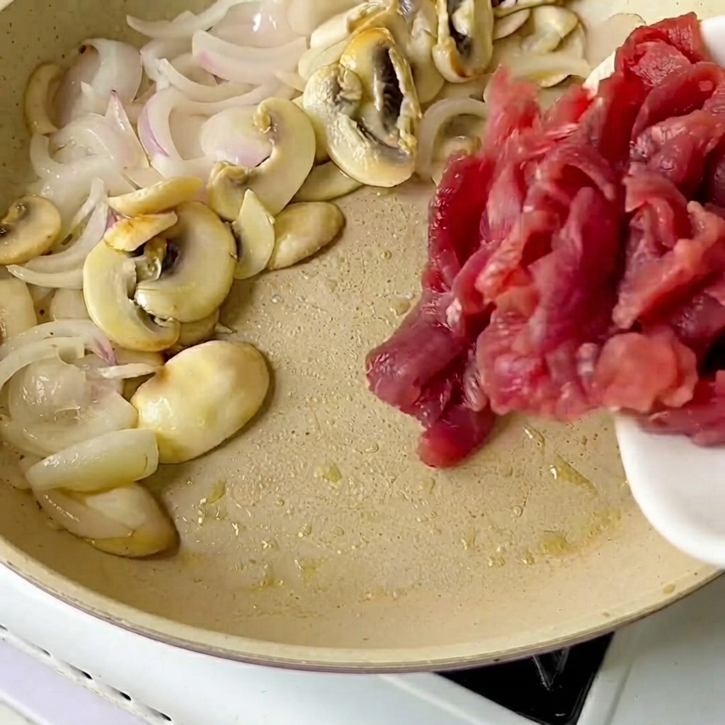 Du bœuf tranché versé dune assiette blanche dans une poêle contenant des oignons et des champignons sautés.
