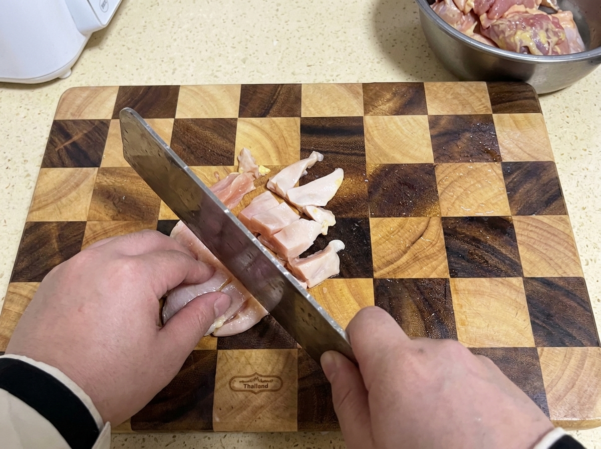 Un couperet coupant de la poitrine de poulet crue en petits morceaux sur une planche à découper en bois à motifs.