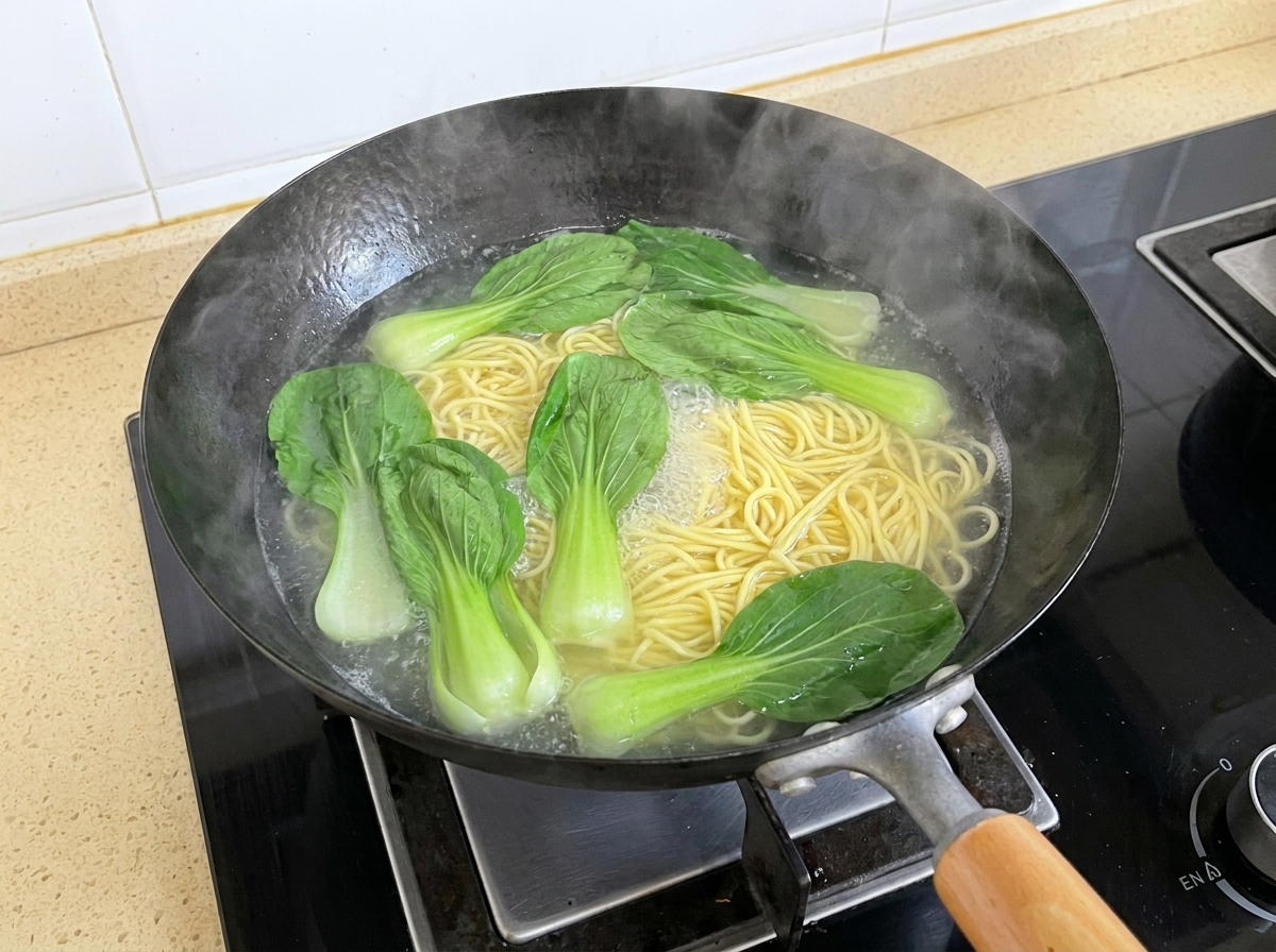 Des feuilles de bok choy fraîches flottant dans leau bouillante aux côtés de nouilles en train de bouillir dans un wok.