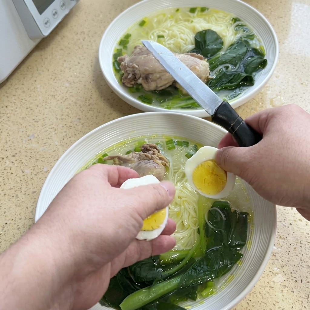 Des mains coupant un œuf dur en deux avec un couteau au-dessus dun bol de nouilles de soupe au poulet.