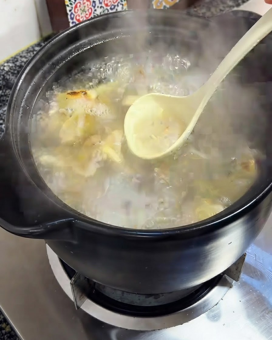 Une louche retirant lécume du bouillon frémissant à lintérieur dune cocotte en terre cuite noire.