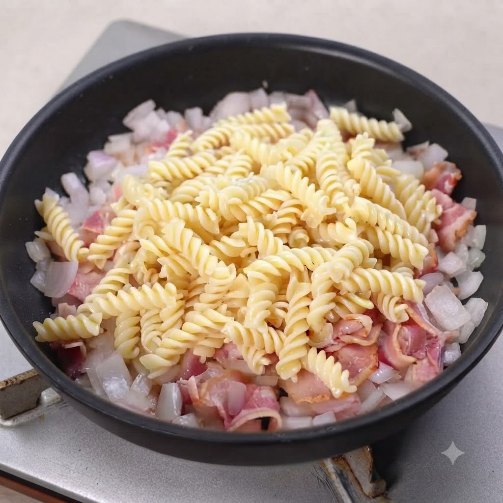 Pâtes spirales cuites reposant sur le bacon et les oignons cuits dans une poêle noire.