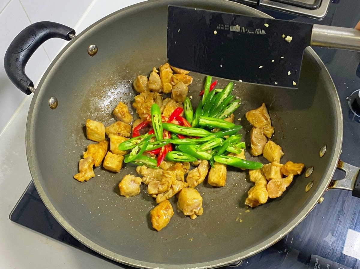 Rondelles de piments verts et rouges ajoutées aux morceaux de poulet cuits dans un wok, mélangées avec une spatule noire.