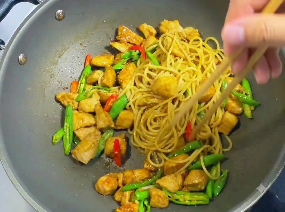 Baguettes en bois mélangeant les spaghetti, les dés de poulet, les rondelles de piments et la sauce de façon homogène dans un wok.