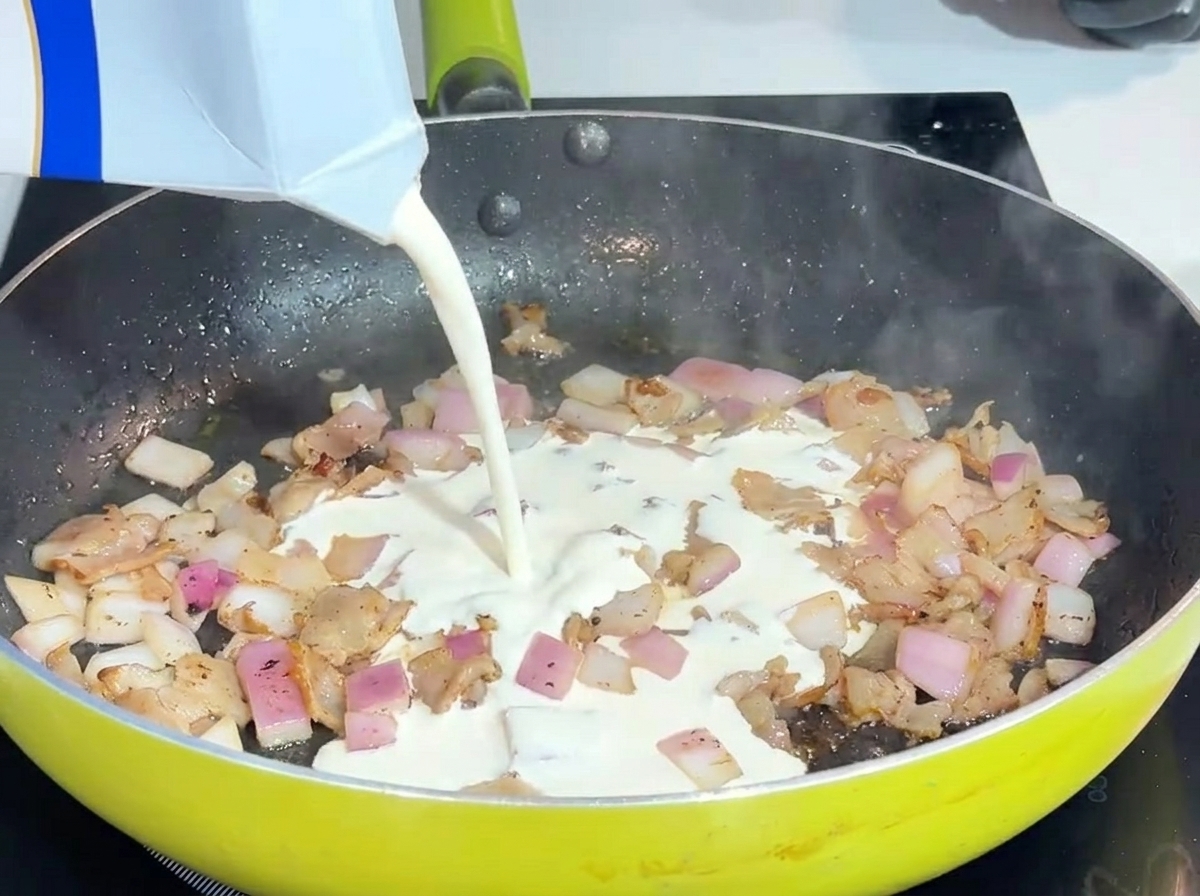 De la crème fraîche liquide versée dans une poêle avec du bacon et des oignons.