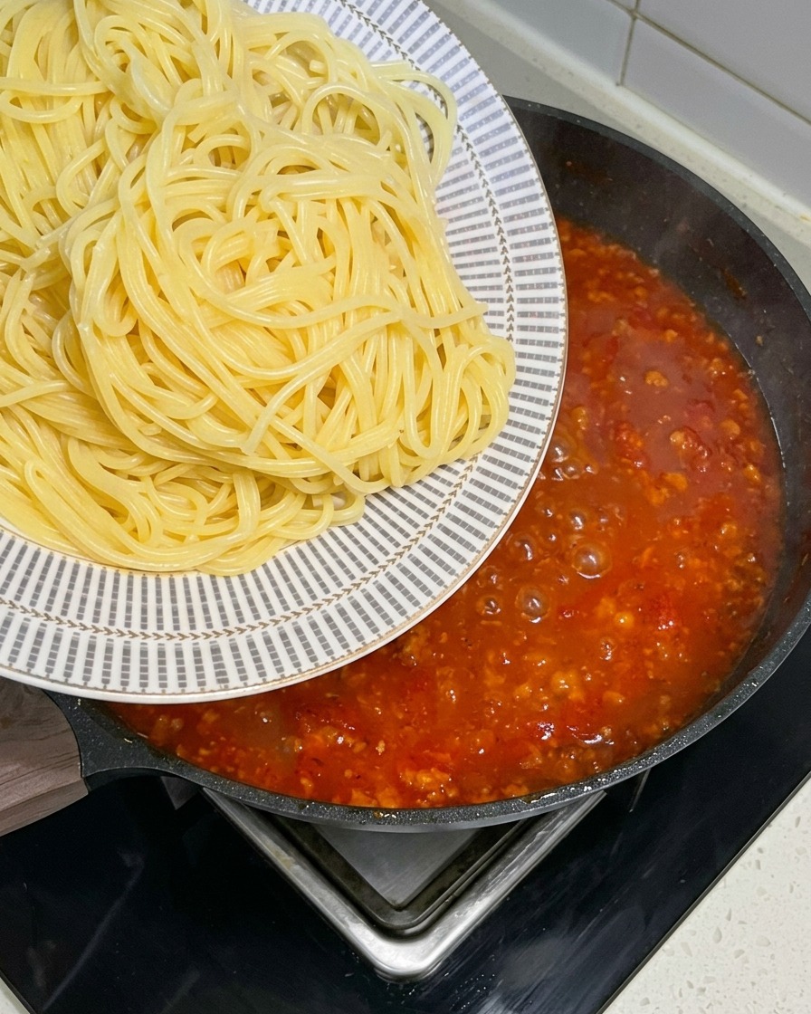 Des spaghettis fraîchement cuits transférés dune assiette à motifs dans une grande poêle remplie dune sauce à la viande frémissante.