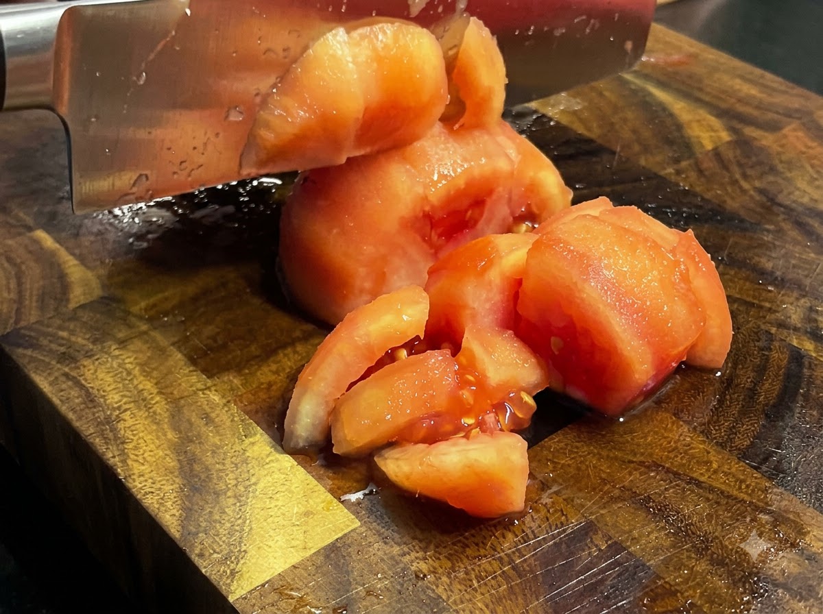 Un couteau de cuisine coupant des tomates cuites et pelées en morceaux sur une planche en bois.