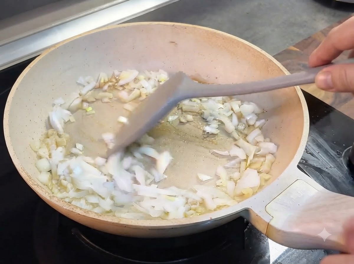 Des oignons hachés remués avec une spatule grise dans une poêle de couleur claire.
