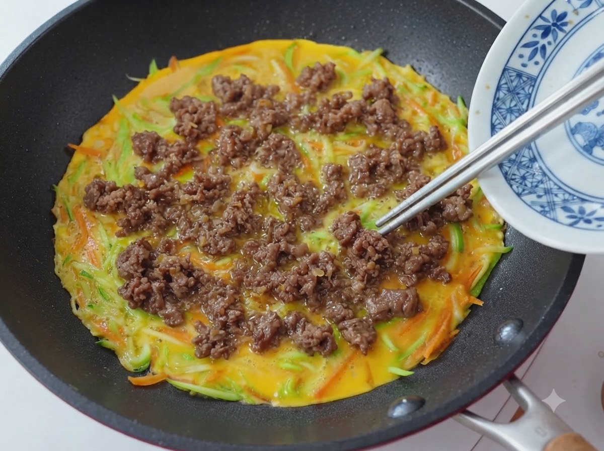 Gros plan de baguettes plaçant et étalant des morceaux de bœuf cuit sur une poêle avec une base de pancake aux légumes et aux œufs.