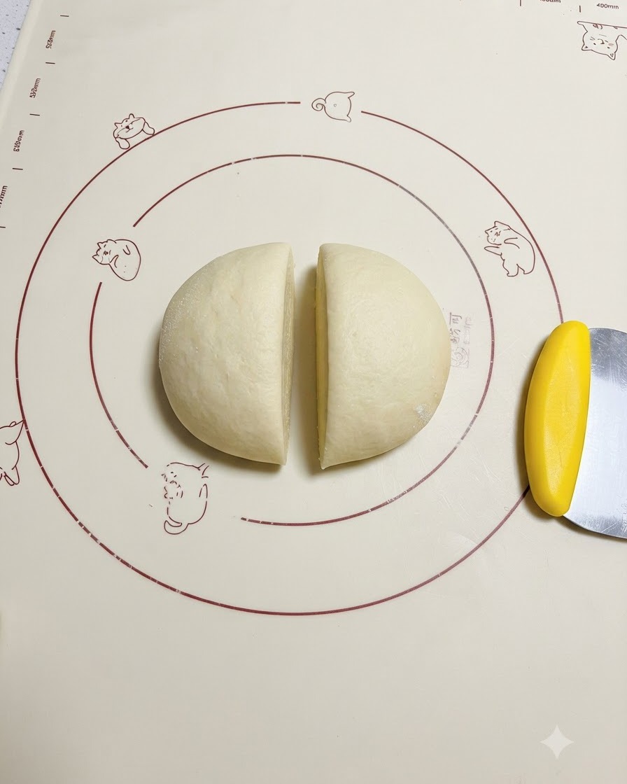 De la pâte levée coupée uniformément en deux sur un tapis en silicone à motifs à côté dun racloir à pâte jaune.