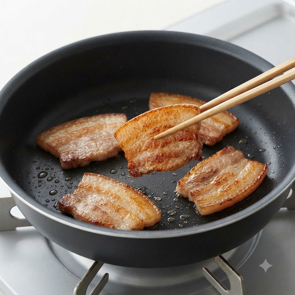 Une paire de baguettes retournant une tranche de poitrine de porc dorée et saisie dans une poêle.