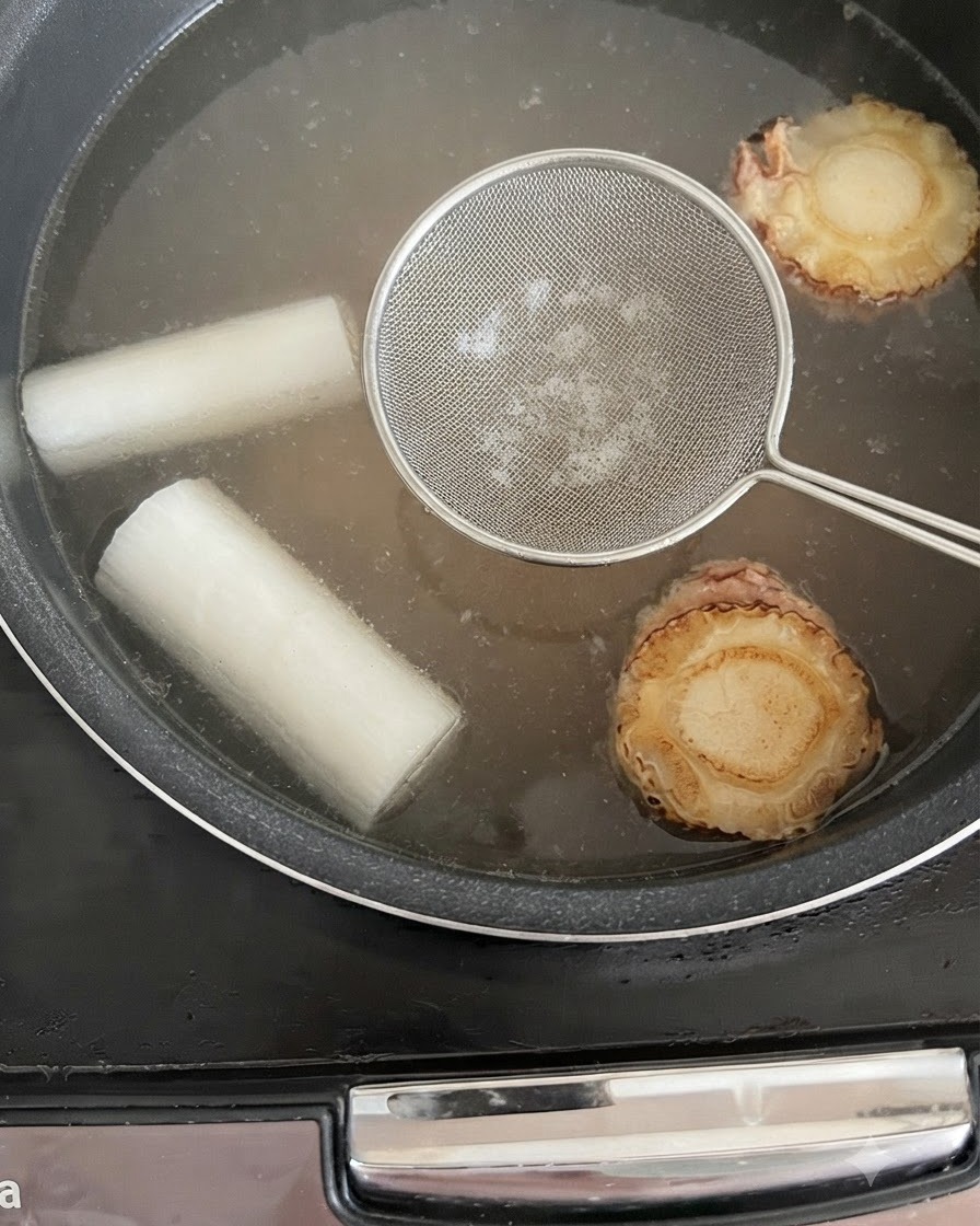 Une écumoire tenue au-dessus du bouillon frémissant dans le cuiseur de riz pour éliminer les impuretés.