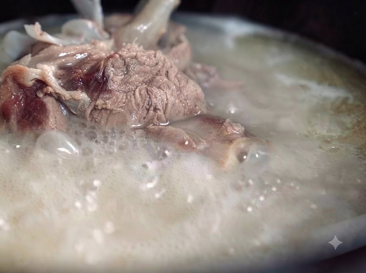 Un bouillon dos de porc epais, laiteux et opaque bouillant vigoureusement dans une casserole, avec des morceaux de viande et dos visibles.