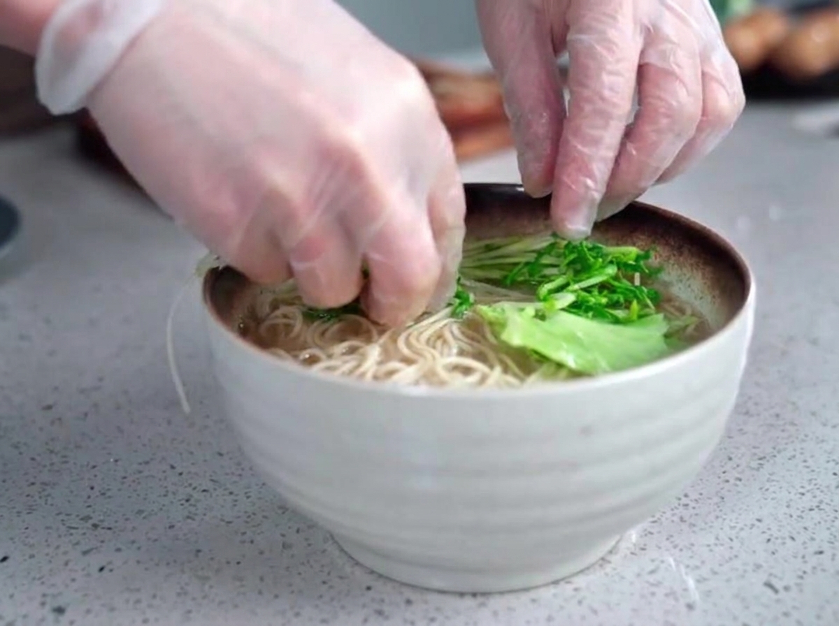 Mains gantees placant soigneusement des legumes verts frais dans un bol fumant de nouilles de ramen et de bouillon opaque.