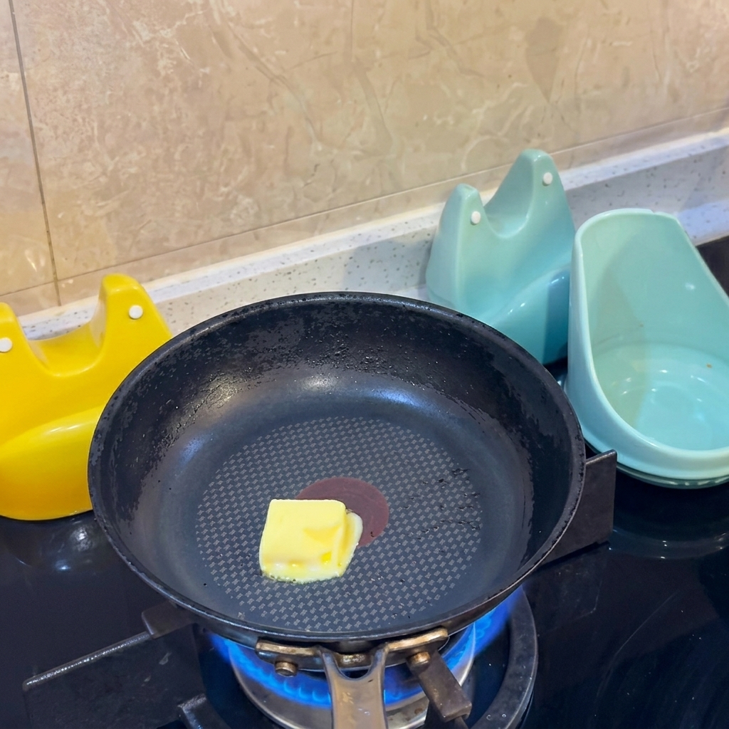Un cube de beurre jaune fonçant au centre dune poêle antiadhésive noire sur une flamme de gaz.