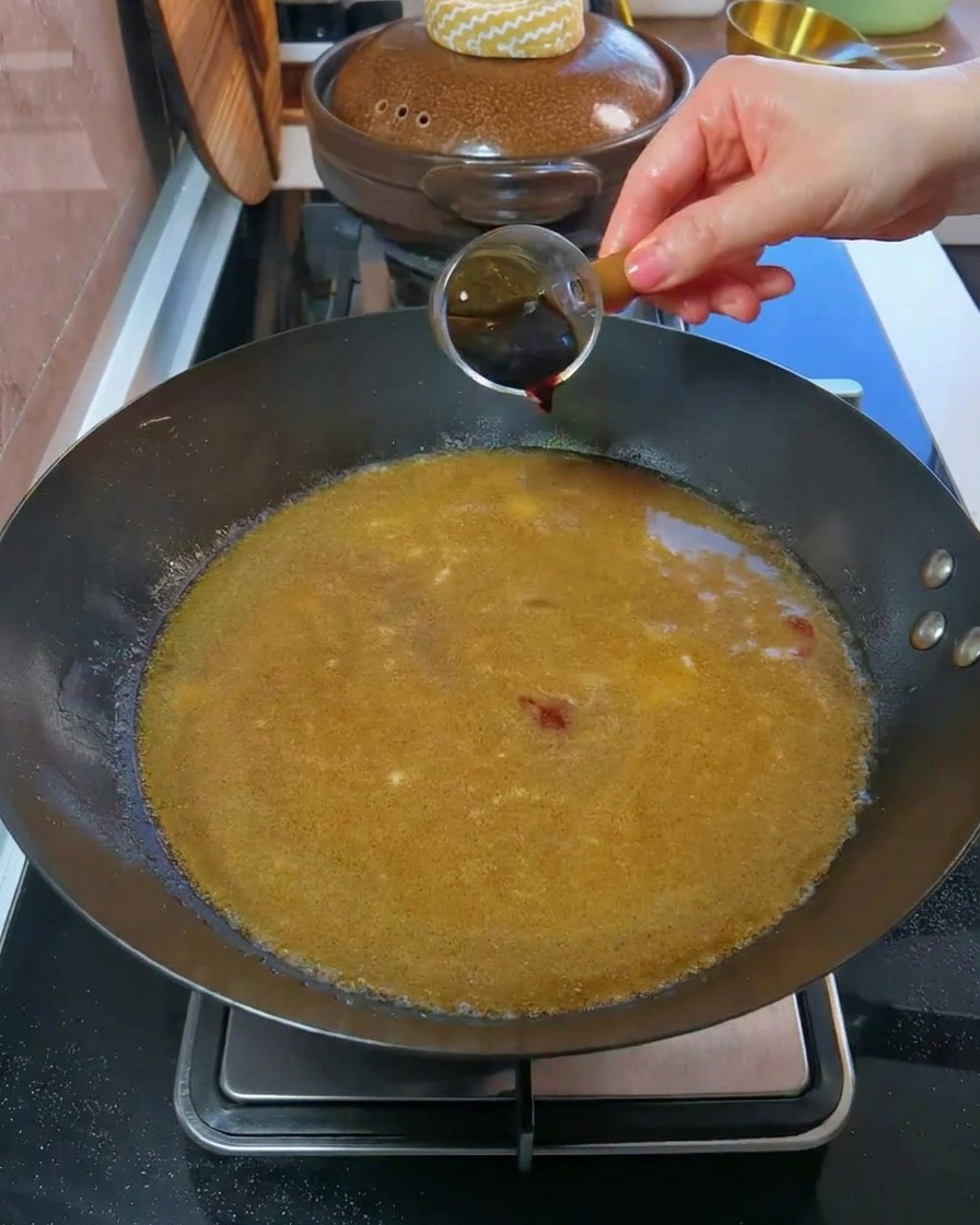 Une main verse de la sauce soja noire dun petit verre doseur dans un wok rempli de bouillon.cite: 1