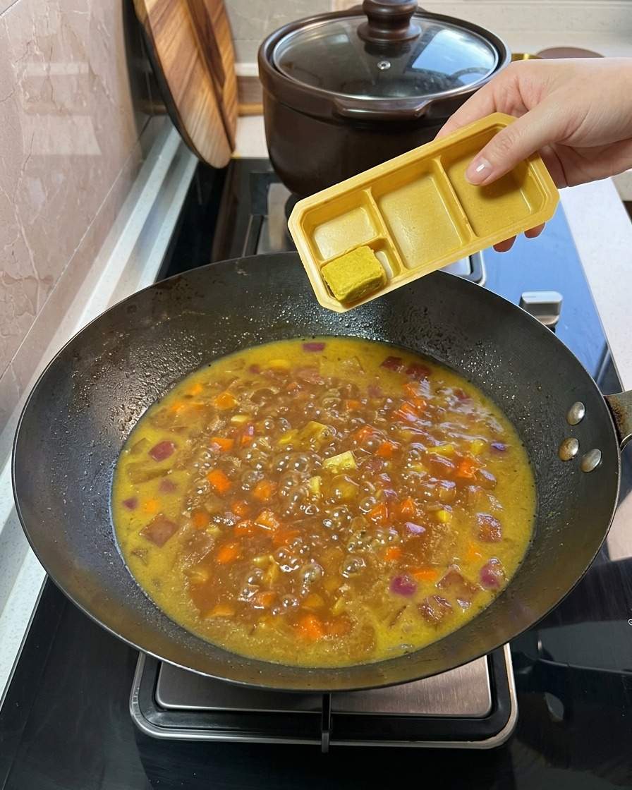 Des cubes de curry solides sont ajoutés dune barquette en plastique dans le mélange de légumes et de bouillon frémissant.cite: 1