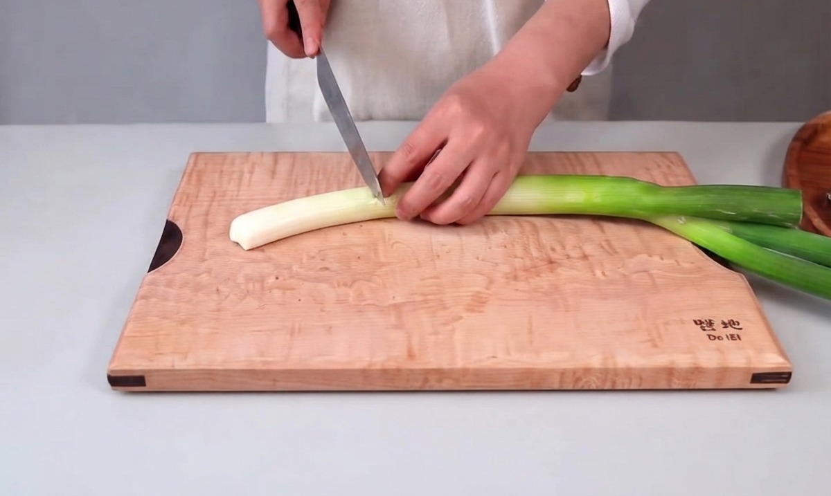 Mains tenant un couteau de cuisine pour émincer une grande ciboule sur une planche à découper en bois.