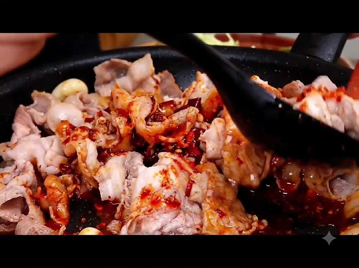 Une spatule noire en train de mélanger et denrober de sauce rouge piquante les tranches de poitrine de porc dans une poêle chaude.