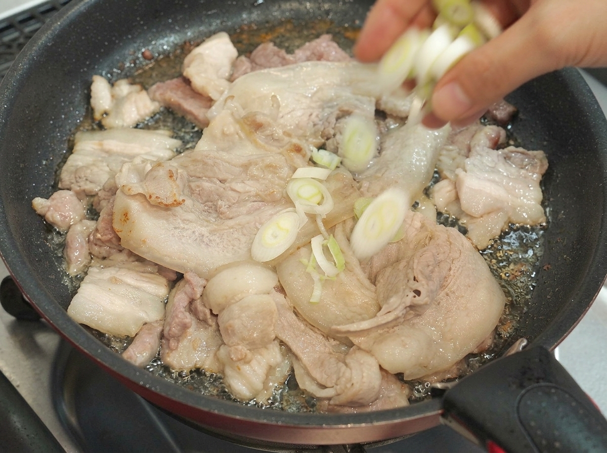 Des oignons verts hachés jetés dans une poêle contenant des tranches de poitrine de porc en train de cuire.