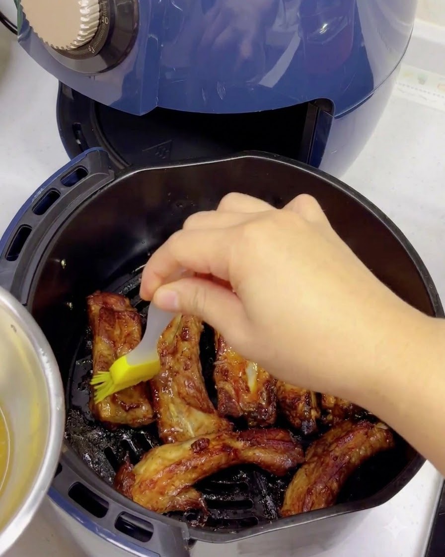 Un pinceau en silicone jaune appliquant un glaçage à leau miellée sur des travers de porc cuits et dorés à lintérieur du panier dun air fryer.