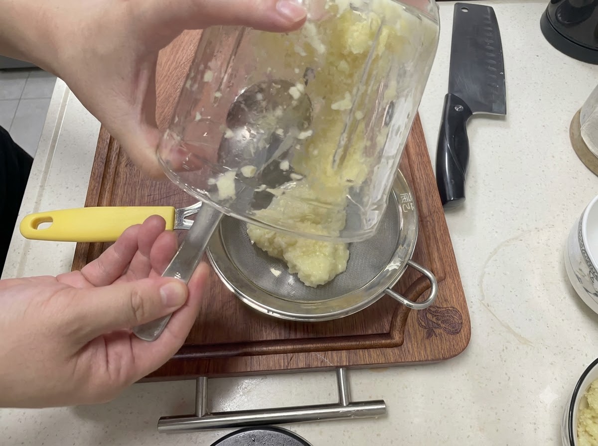 Ail mixé versé dun récipient en verre dans une passoire à mailles fines au-dessus dun bol en métal.